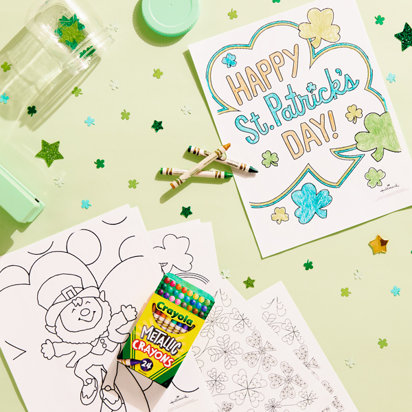 St. Patrick's Day coloring pages spread out on a light green surface, surrounded by crayons in different shades of green and green and gold star-shaped confetti.