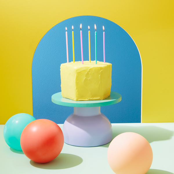 A yellow birthday cake with tall slim candles sitting on a pedestal and surrounded by colorful balloons.