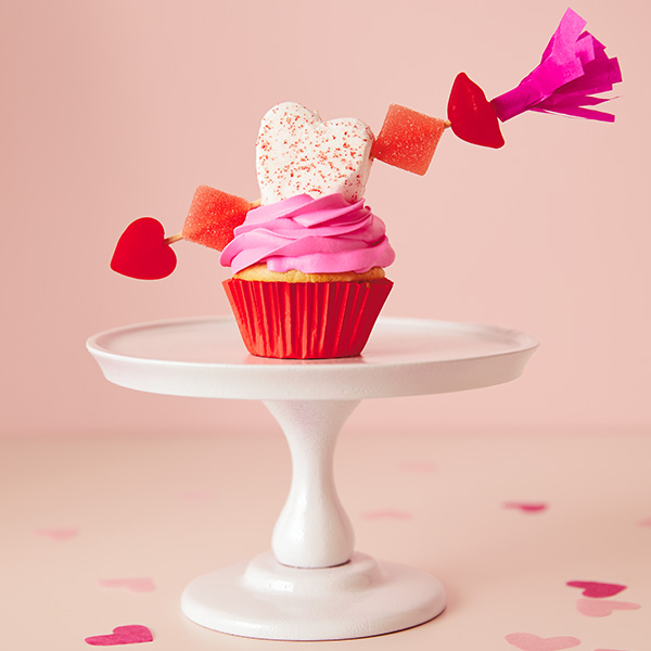 Valentine's Day cupcake with pink frosting, a marshmallow heart on top and an arrow garnish made of gummy candy.