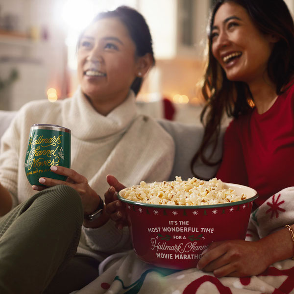 Two women sitting on a couch watching TV; one holding green stemless wine glass, other holding red popcorn bowl