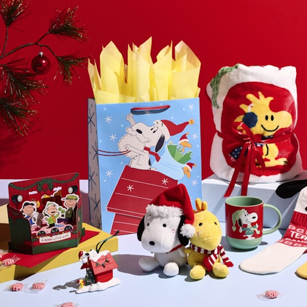 A collection of Peanuts-themed Christmas gifts and cards sits on a table against a cheery red background with a spindly little evergreen tree decorated with a single red glass ball ornament, as in the classic Charlie Brown Christmas movie.