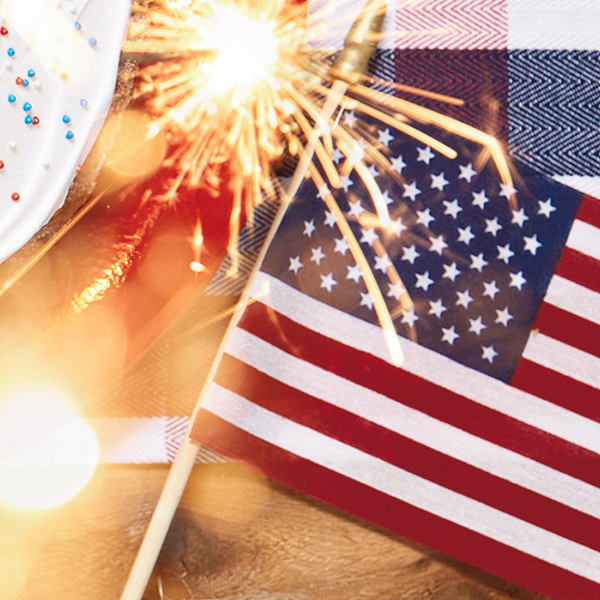 An American flag on a tabletop with a red, white and blue table runner, next to a cake decorated with red, white and blue sprinkles and a lit sparkler firework.