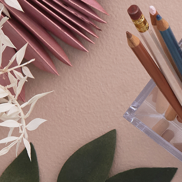 A clear acrylic pencil cup with multicolored pencils in it, with various leaves and paper textures nearby.