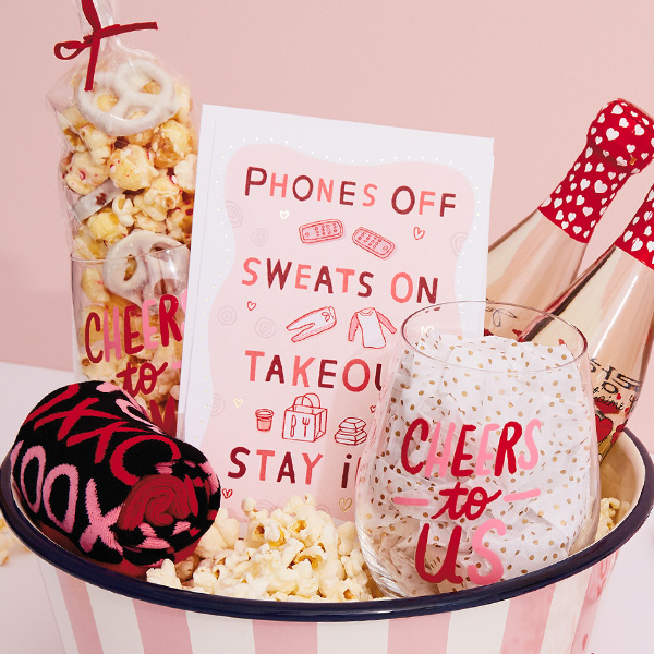 A Valentine's Day care package filled with gifts for a movie night in, including a Hallmark Channel popcorn bowl, wine glasses, cozy socks and a card.