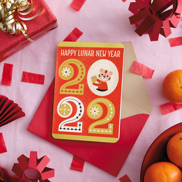 A 2022 Lunar New Year card on a table surrounded by red paper lanterns, red confetti, mandarin oranges, gifts and fans.