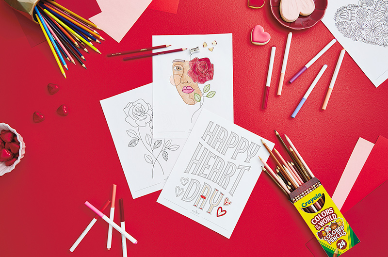 A partially colored in free printable Valentine's Day coloring page on a red table top scattered with Crayola colored pencils and markers.