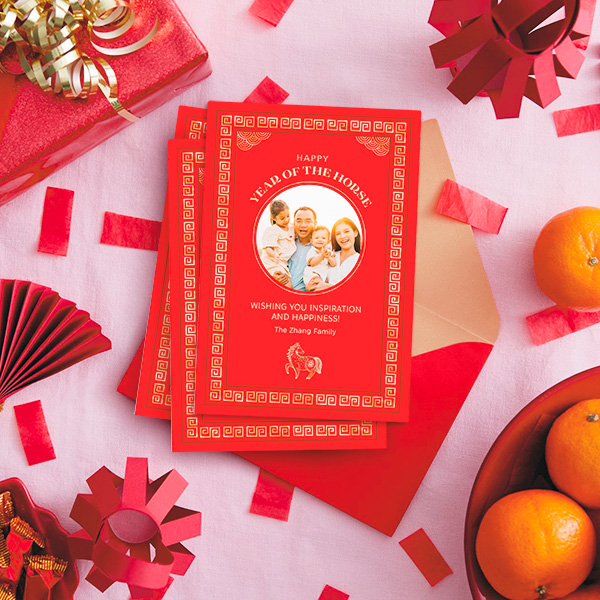A customizable Lunar New Year card featuring a family photograph sits on top of a red envelope, on a table covered in red paper lanterns and red confetti; a bowl of oranges sits nearby.