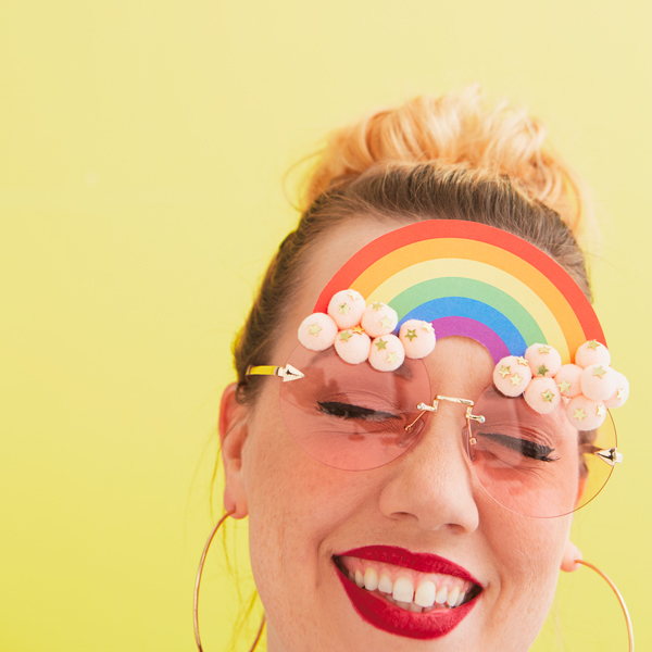 A woman wearing bright red lipstick and fun Pride Month rainbow sunglasses, smiling with her eyes closed.