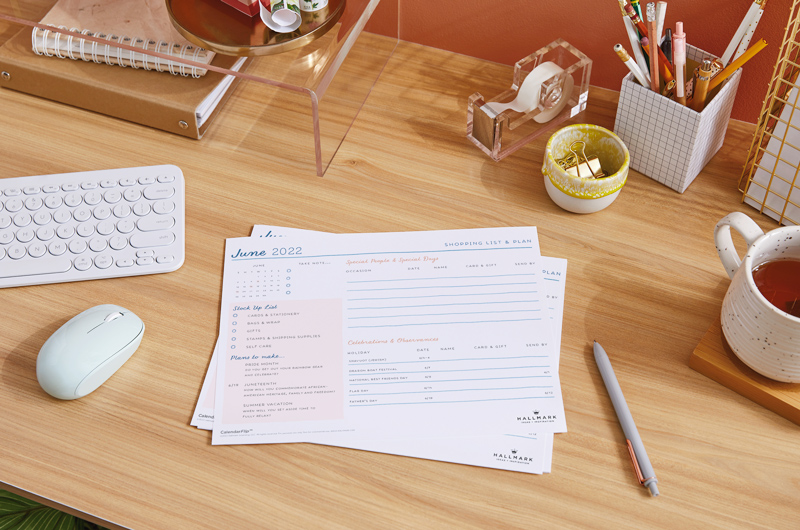 A desktop surface with a keyboard, mouse, a mug of tea and office supplies sitting on it; a stack of celebration planner pages sits at the center, with a page for the month of June on top.