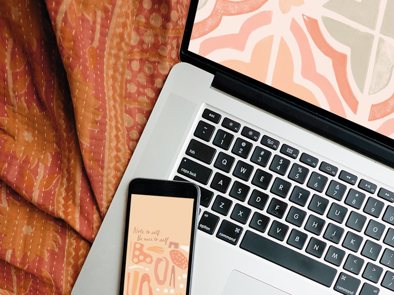 A laptop and a smartphone sit on a red and orange patterned blanket; both of their screens feature a digital wallpaper.