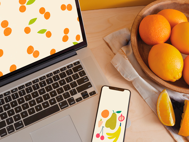 A laptop and a smartphone on a wooden countertop, both displaying digital wallpapers; a bowl of oranges sits nearby.