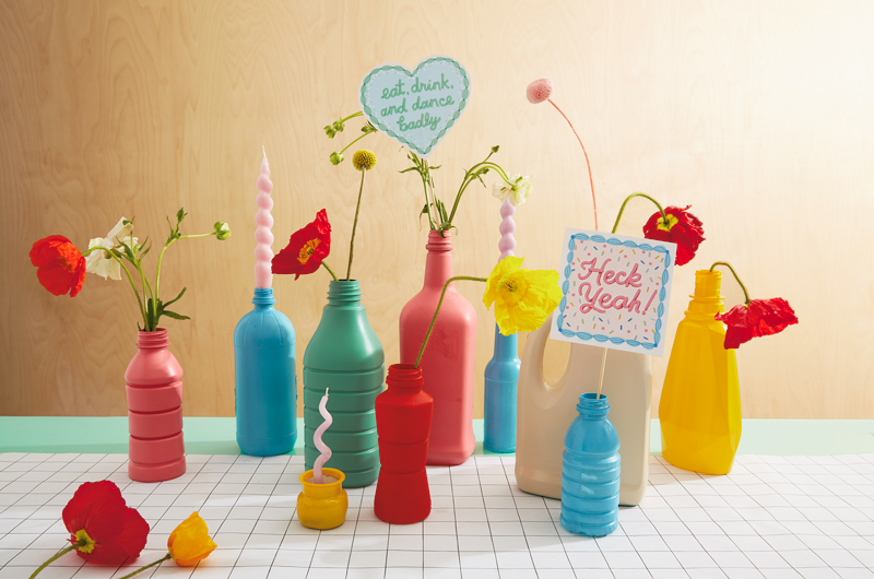 A group of repurposed plastic bottles spray painted in bright colors and serving as vases on a tabletop with a black and white checkered surface.