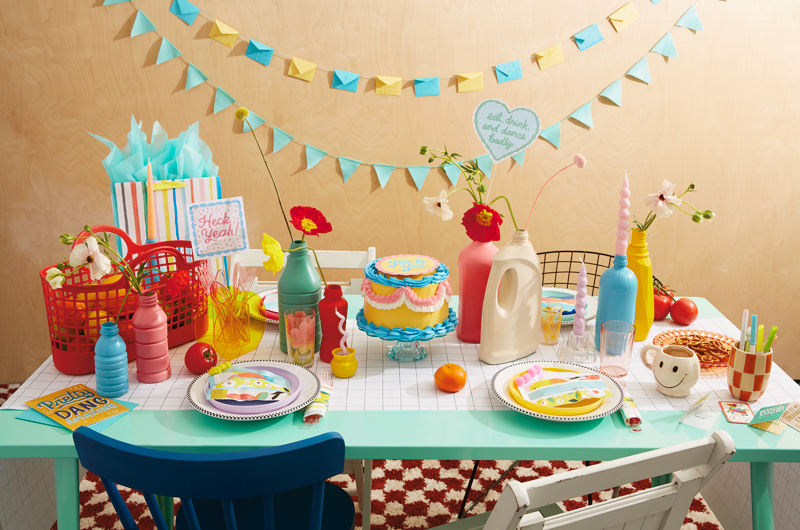 A table set with DIY party decorations, including a cake with a free printable cake topper, a mini card envelope garland, and cake favor pouches.