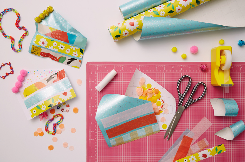 Materials used to create the cake slice favor pouches scattered across a work surface, including wrapping paper, tape, scissors, mini pom-poms and glue.