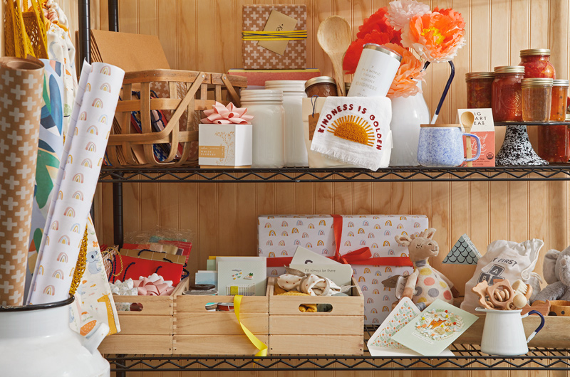 A gift closet setup with homey gifts like homemade preserves, kitchen towels, tumblers and wooden utensils, as well as a selection of baby gifts and gift wrap in muted, natural colors.