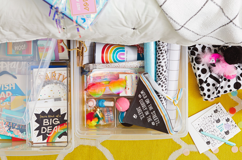 Two clear storage bins sticking out from under a bed, filled with colorful cards and small gifts like nail polish, pet bandanas and toys, tumblers and friendship bracelets.