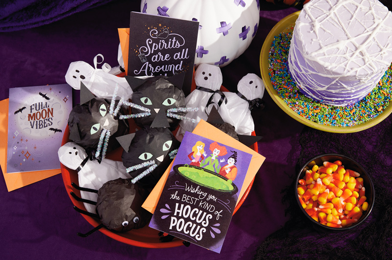 A Hocus Pocus party treat table with a spiderweb cake, black cat and ghost tissue paper treats, Halloween greeting cards, and candy corn.