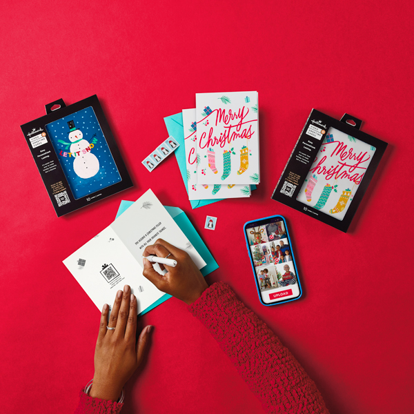 A woman's hands hold open a Hallmark Holiday Video Greeting card to write in it, while a box with more cards and a smartphone with the Video Greetings interface showing on its screen lay nearby.
