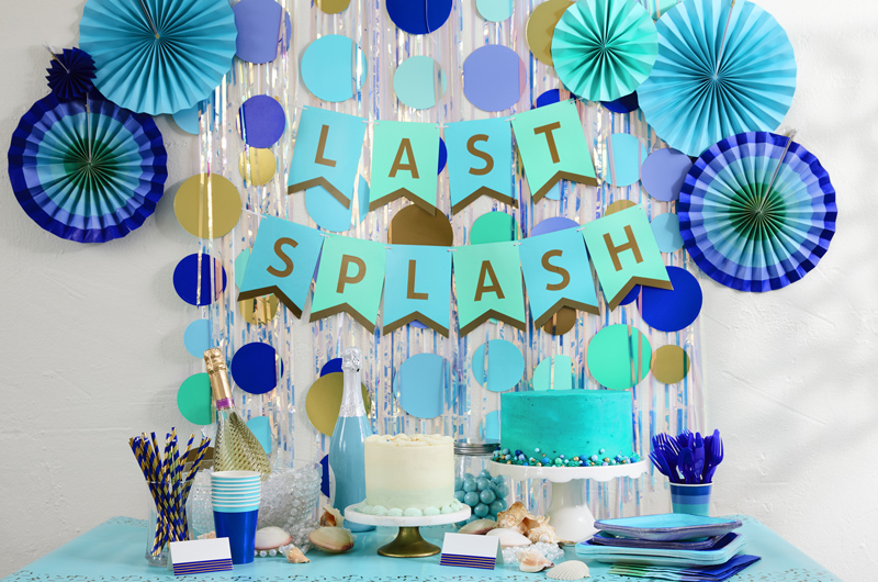 A summer beach or pool party table with two cakes surrounded by Color Pop party ware in shades of blue. Behind the party table is a banner that reads 