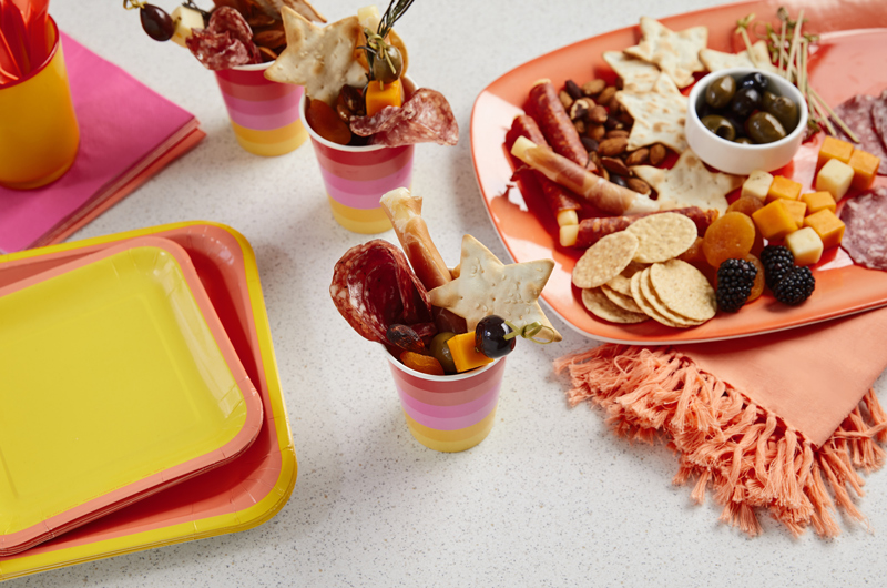 A red, pink and yellow ombre Color Pop party ware cup holds slices of meat, cubes of cheese and crackers, functioning as a single-serve charcuterie cup or 