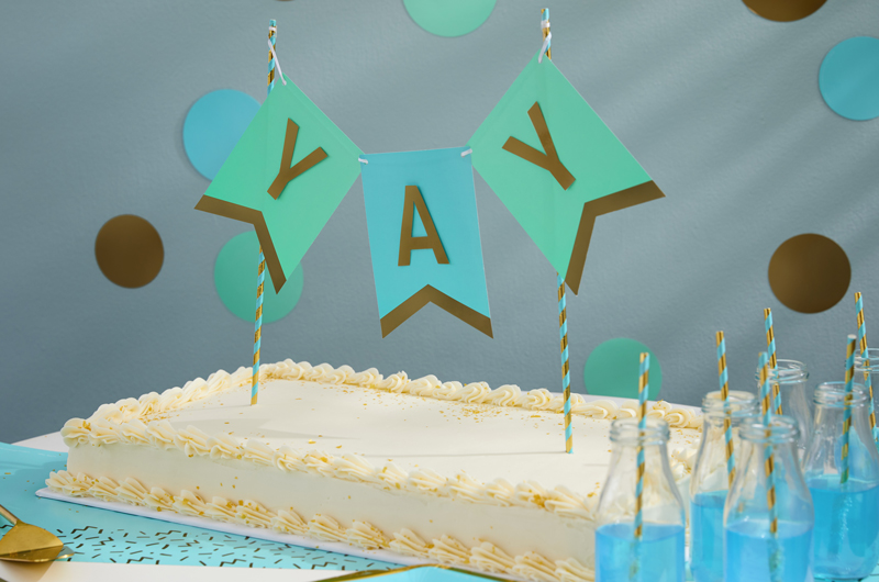 A DIY birthday banner cake topper inserted into a white sheet cake piped with frosting. Oversized confetti is adhered to the wall behind the cake.