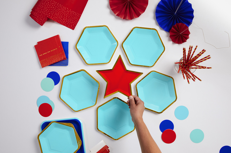 A hand places a red plate in the shape of a star in the center of a patriotic serving set up, featuring light blue and gold hexagon-shaped plates and red and dark blue paper fans.