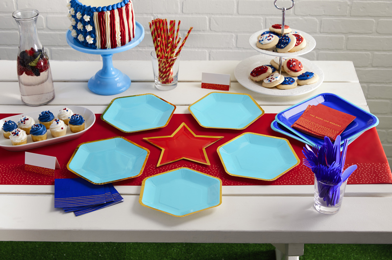 A patriotic party table, featuring light blue and gold hexagon-shaped plates, a red star-shaped plate, and red and dark blue paper fans.