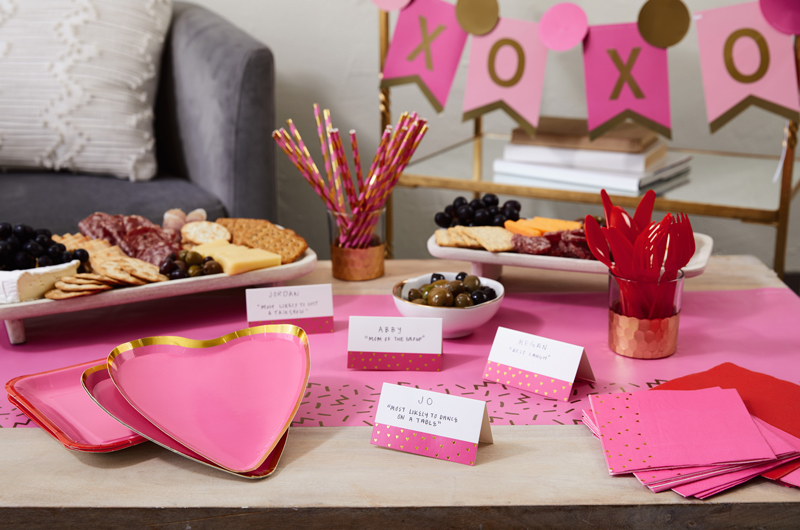 A Galentine's Day party featuring pink and gold Color Pop heart-shaped plates, pink and gold striped paper straws, and a pink and gold banner that reads 