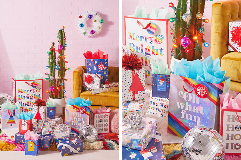 A large number of presents wrapped in bright, rainbow-colored and holographic gift wrap and bags. In the background is a cactus wrapped with Christmas lights and ornament bulbs.