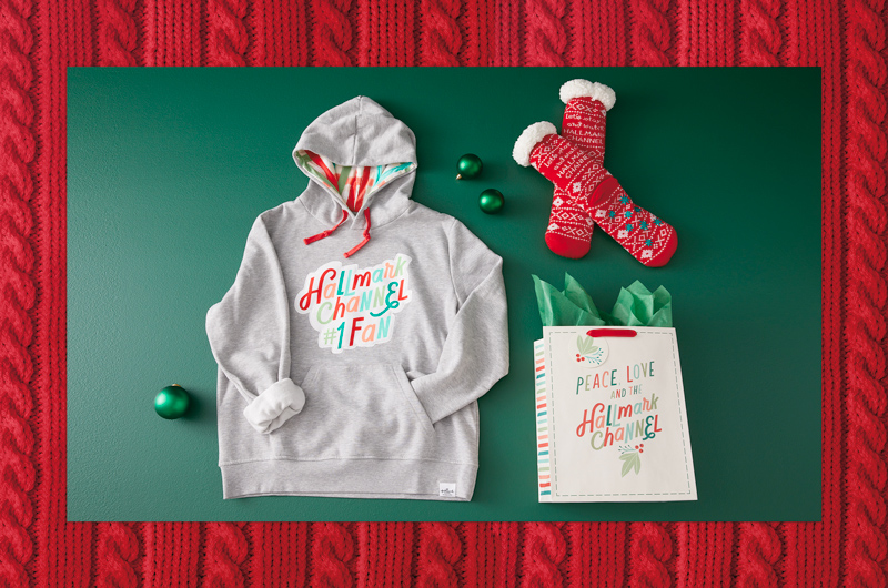 Gift ideas for the Hallmark Channel newbie in your life include a Hallmark Channel #1 Fan hoodie, and a plush pair of cozy Hallmark Channel slipper socks, wrapped up in a gift bag that reads, 