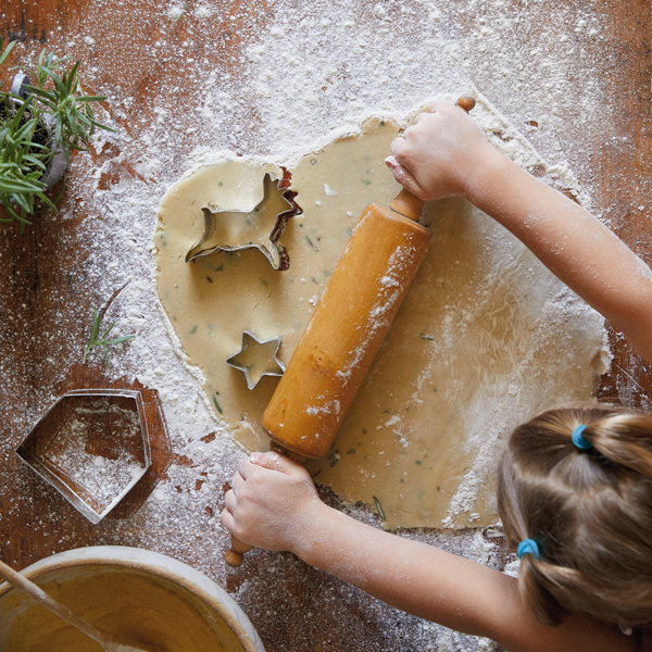 A little girl rolls out cookie dough with a rolling pin on a floured surface, with different shapes of cookie cutters nearby.