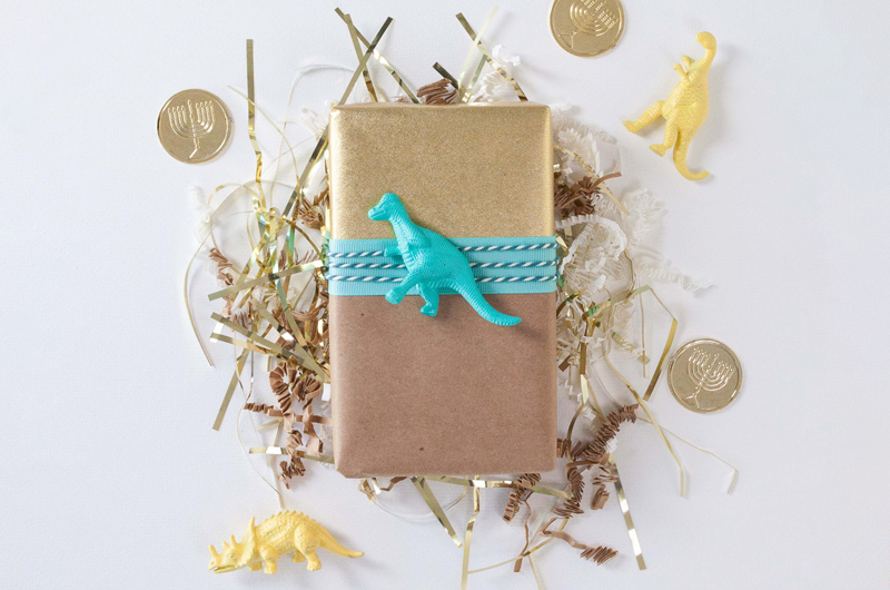 A small gift wrapped in gold and brown Kraft paper wrapping, decorated with blue and white cotton cording and a blue plastic toy dinosaur.