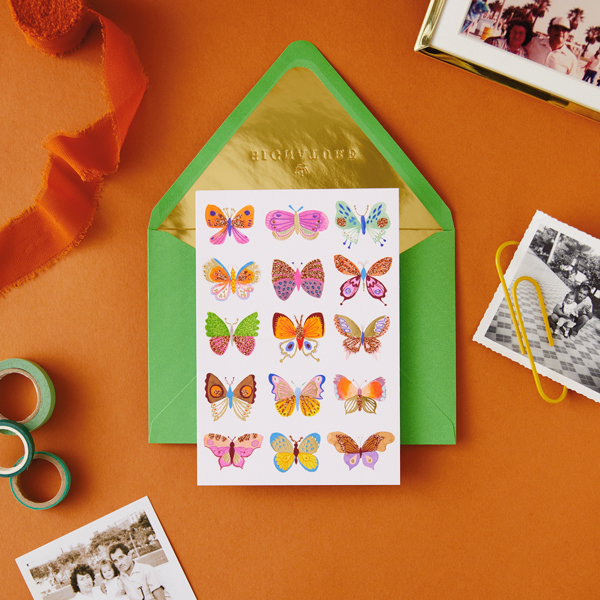 A card featuring several butterflies of different colors and patterns, lined up in three columns on the front. Lying under the card is a bright green envelope with gold foil lining inside.