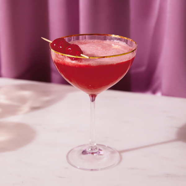 A martini-style cocktail glass with a gold rim, filled with a red cocktail topped with a pink egg white foam and garnished with maraschino cherries on a cocktail pick.