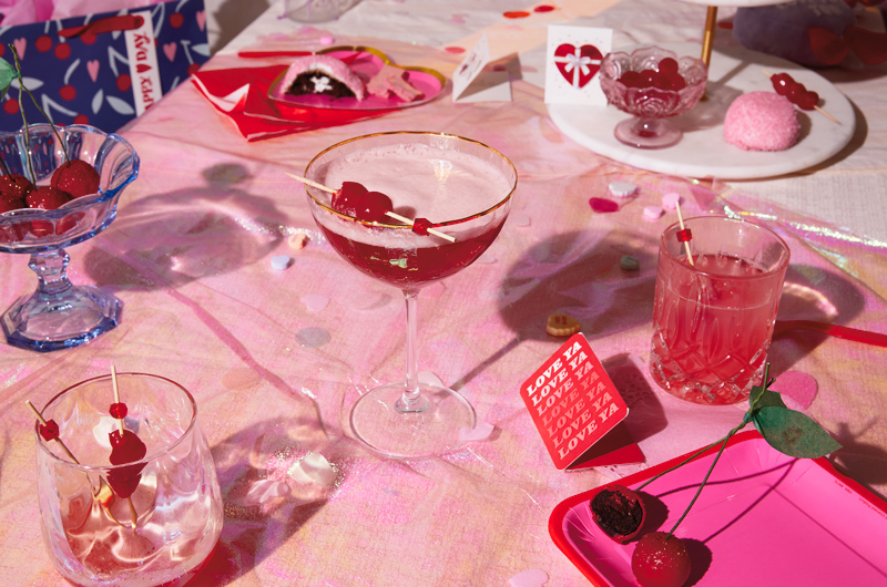 A martini-style cocktail glass with a gold rim, filled with a red cocktail topped with a pink egg white foam and garnished with maraschino cherries on a cocktail pick sits on a table amid the remnants of a Valentine's Day party—plates, napkins, additional cocktail glasses, cards and gifts.