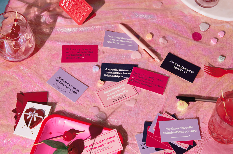 Conversation Starter cards are scattered atop a table set for a Valentine's Day cocktail party and strewn with candy conversation hearts, cocktail glasses and plates with cake truffles shaped like cherries.