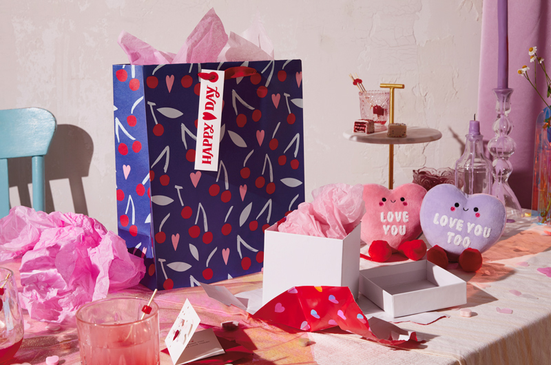 A gift bag featuring a pattern of cherries and hearts on a dark blue background sits on a table decorated for a Valentine's Day party, along with a set of conversation heart magnetic plushes.