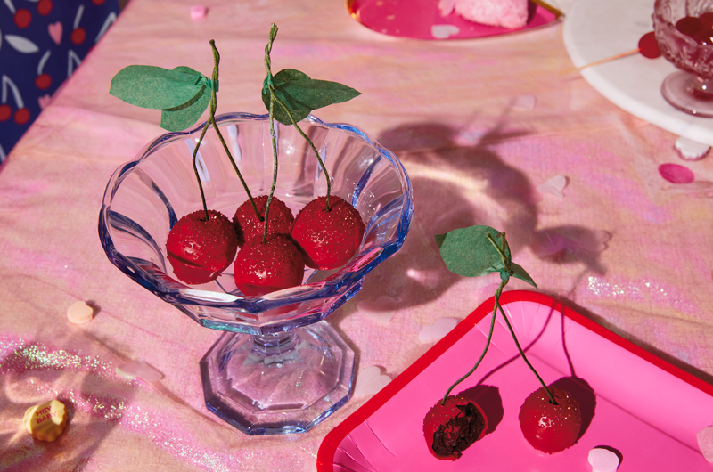 Chocolate cherry cake truffles decorated to look like real cherries sit in a clear blue glass dessert dish with a scalloped rim on a table scattered with the remnants of a Valentine's Day party.