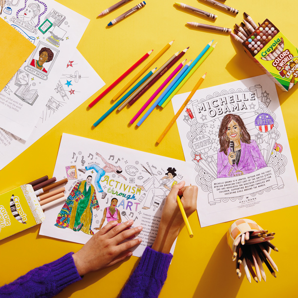 A child's hands color in a Black History Month coloring page. Scattered around the bright yellow work surface are crayons and colored pencils in various colors.