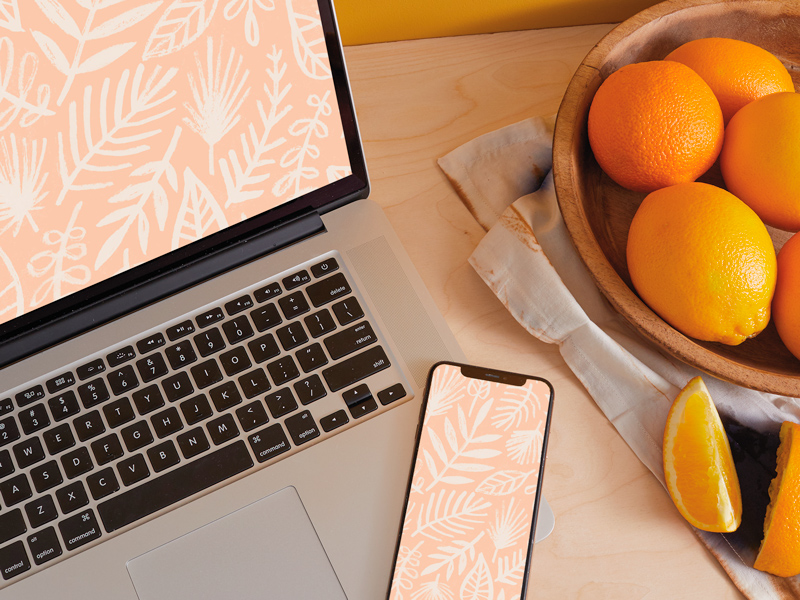 A laptop displaying a digital wallpaper featuring white botanical illustrations against a light orange background sits on a desktop next to a mobile phone displaying the same background on its screen.