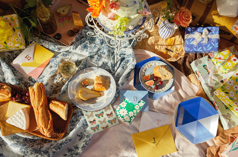 Plates of fruit, cheese and bread, cards with cross-stitch monogrammed envelopes, gifts and a cake stand with a cake and butterfly cake topper are arranged on the floor for an indoor spring picnic party.
