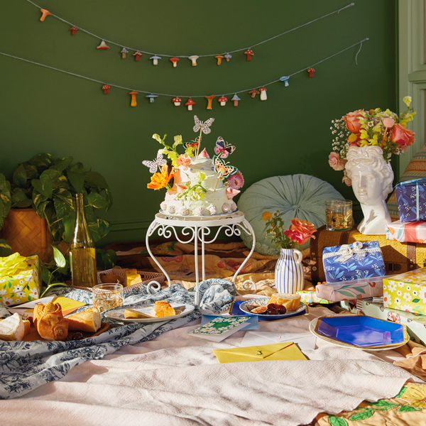Plates of fruit, cheese and bread, cards with cross-stitch monogrammed envelopes, gifts and a cake stand with a cake and butterfly cake topper are arranged on the floor for an indoor spring picnic party. A DIY mushroom garland hangs in the background.