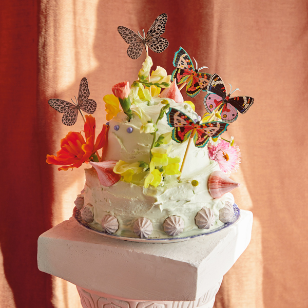 A cake decorated with meringues, edible pearls and edible flowers, and a DIY butterfly cake topper sit atop a cake stand that looks like a Roman column.