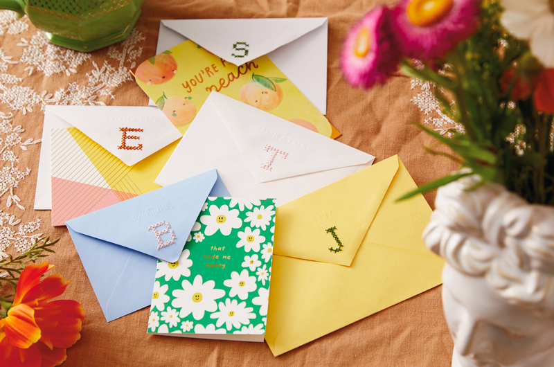 Five envelopes, each with a different initial monogrammed in cross-stitch onto the back flap, along with a selection of Hallmark cards.