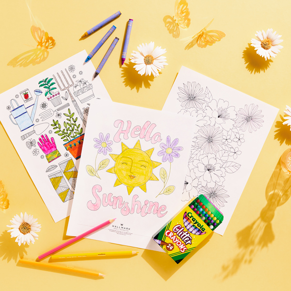 Free printable spring coloring pages spread on a light yellow surface and surrounded by a scattering of crayons, colored pencils and daisies.