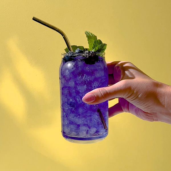 A woman's hand holding a glass filled with bright purple sparkling butterfly pea tea, garnished with mint and berries.