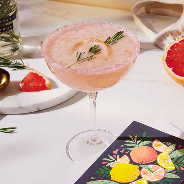 A pink cocktail in a coupe glass with a sugared rim and garnished with rosemary sits on a table with a Signature card that features an illustration of citrus fruit on the front.