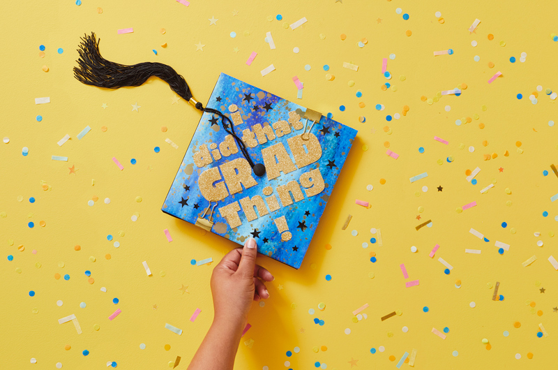 A DIY graduation cap decoration idea—the graduation cap is covered with a piece of chipboard that's wrapped in Hallmark wrapping paper and has gold, glittery letters on it that read, 