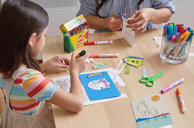 A little girl and a woman sit at a table coloring free printable Father's Day coloring pages and cutting out badges and banners to paste onto his Father's Day card envelope.