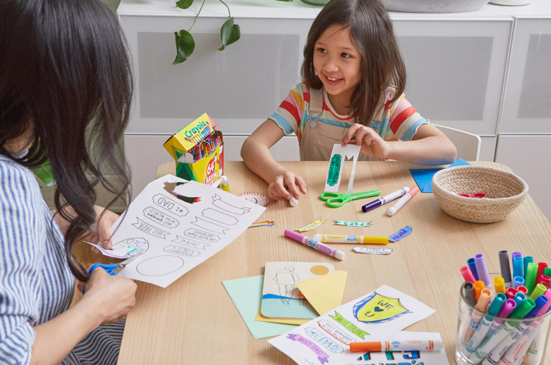 A little girl holds up a piece of a free printable Father's Day banner and badge coloring page as her mom cuts another out of a piece of paper.